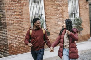 Young satisfied multiethnic partners speaking while looking at each other and strolling against brick building in town